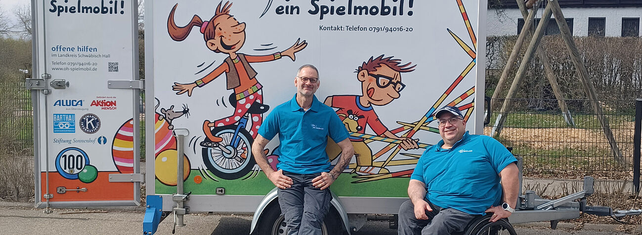 Zwei Mitarbeitende vor einem bunt gestalteten Spielmobil-Anhänger